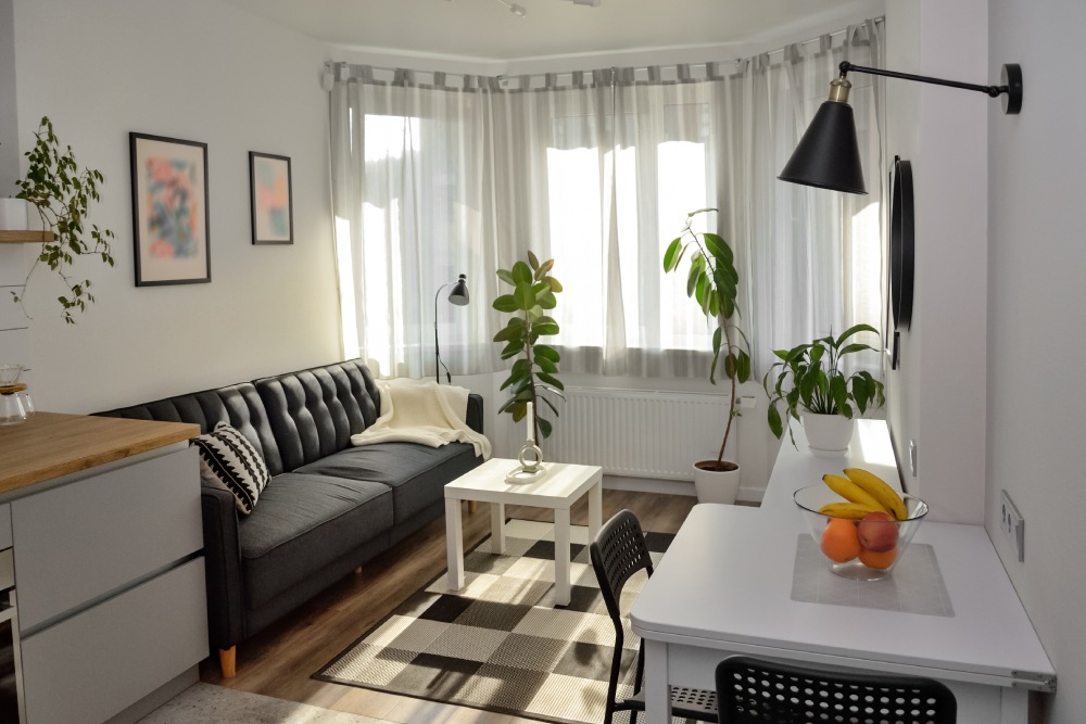 Büyük pencerelerden ışık alan salon içerisinde siyah koltuk beyaz yemek masası ile 1+1 Ev Dekorasyonu önerileri