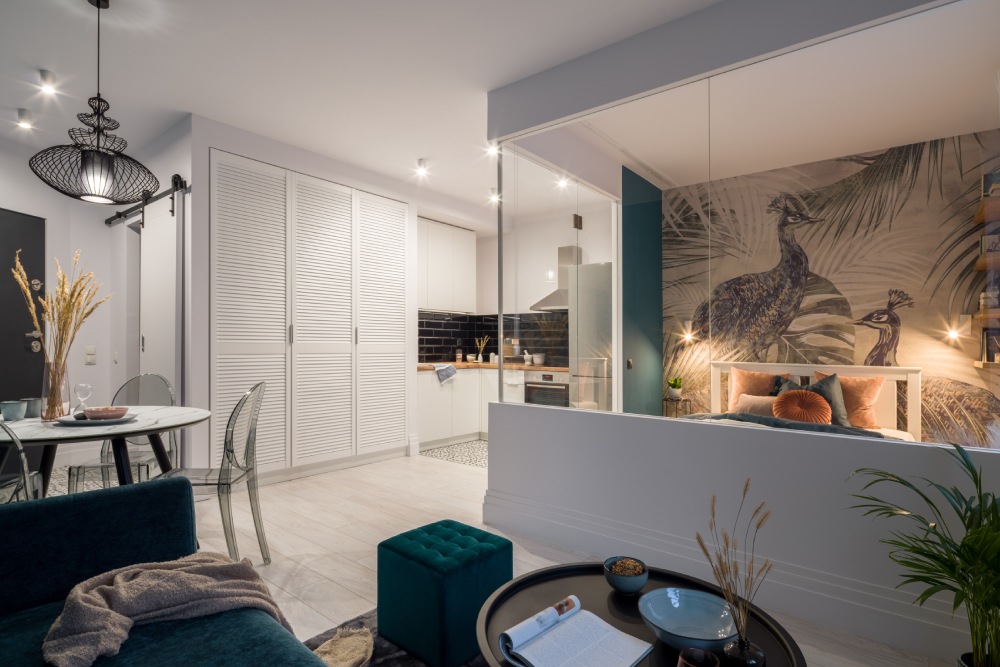 Oturma odası, yemek masası ve mutfağın aynı alanda yer aldığı; cam duvarın arkasında aydınlatılmış yatak odası bulunan, küçük ama şık tasarlanmış 1+1 Ev dekorasyonu.