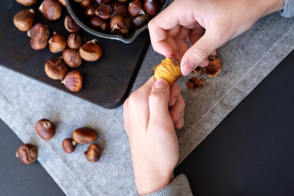 Fırında kestane yapımı ipuçları. Mutfak masasında kestanelerle dolu koyu ahşap bir tahta ve keten masa örtüsü üzerinde kavrulmuş bir kestaneyi soyuyor. 